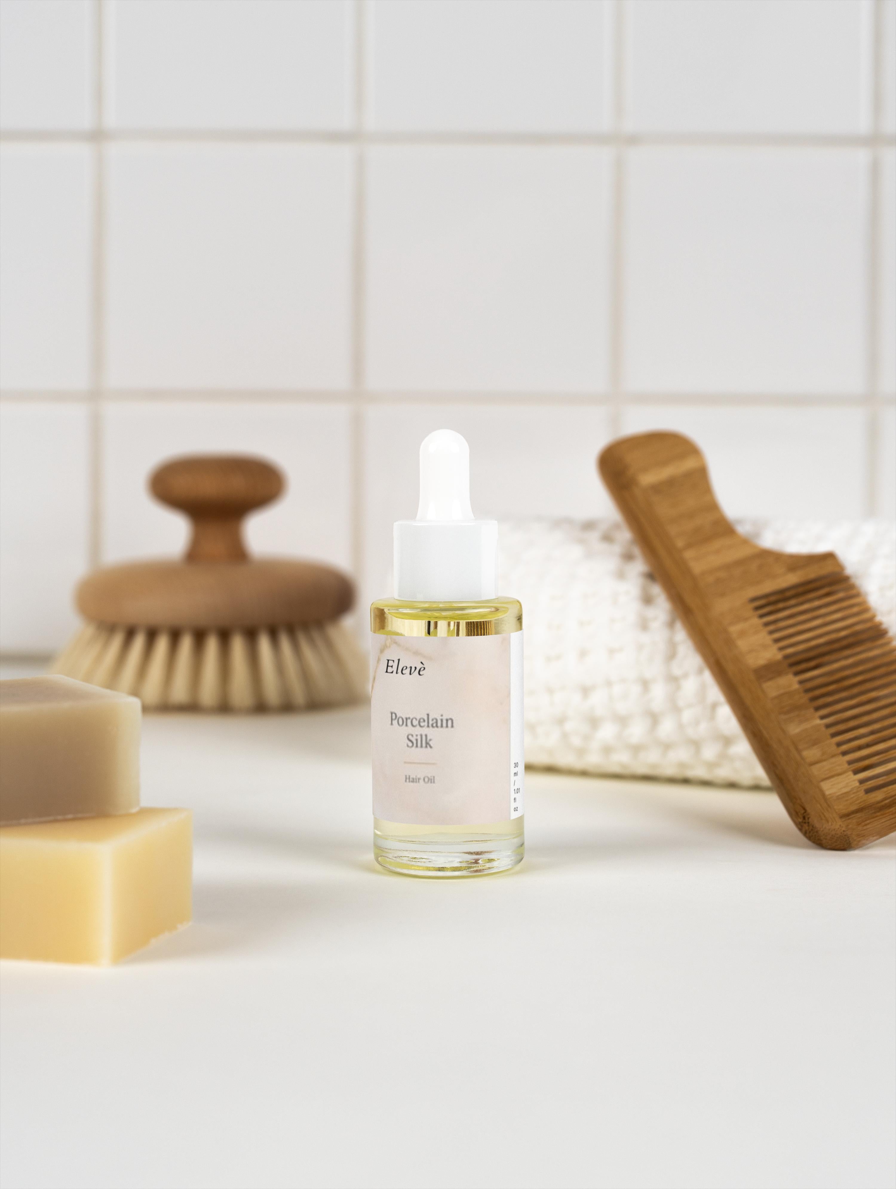 Elegantes Elevè Haaröl im Glasflakon auf weißer Oberfläche im Badezimmer, neben Kamm und Seife, für gepflegtes und glänzendes Haar